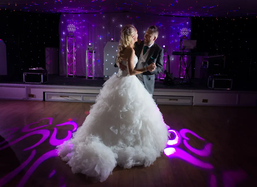 Bride in a voluminous white gown and groom in a suit share a dance on a lit wooden floor with purple spotlight patterns.