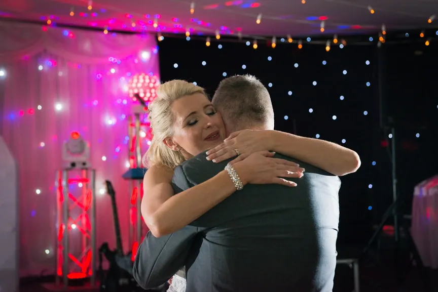 Bride and groom warmly embracing during their wedding dance with colorful lights and stage equipment in the background.