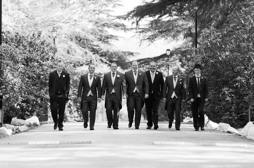 Seven men in formal suits walking together down a tree-lined path outdoors.