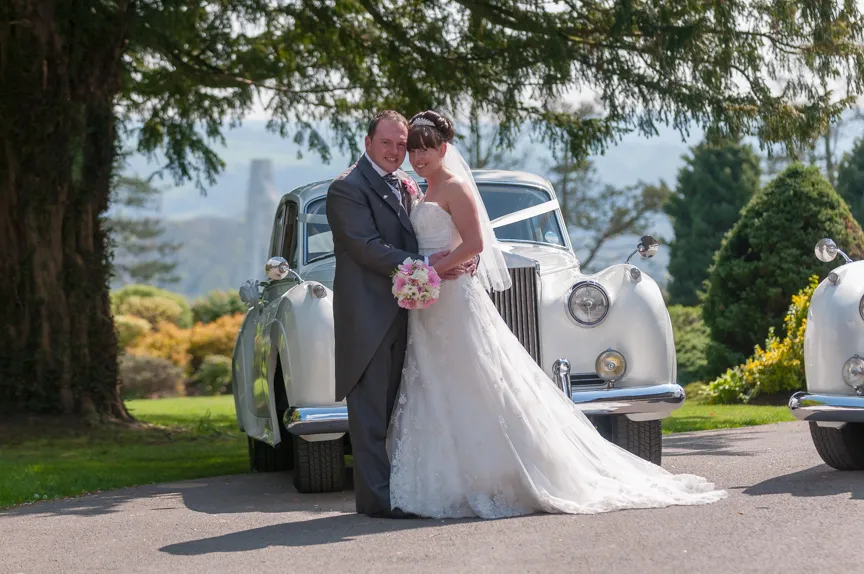 Bride and groom embracing in front of a vintage white car decorated with wedding ribbons on a sunny day.