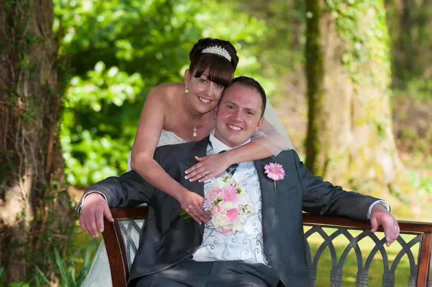 Bride in white dress hugging groom in dark suit sitting on wooden bench outdoors with greenery background.