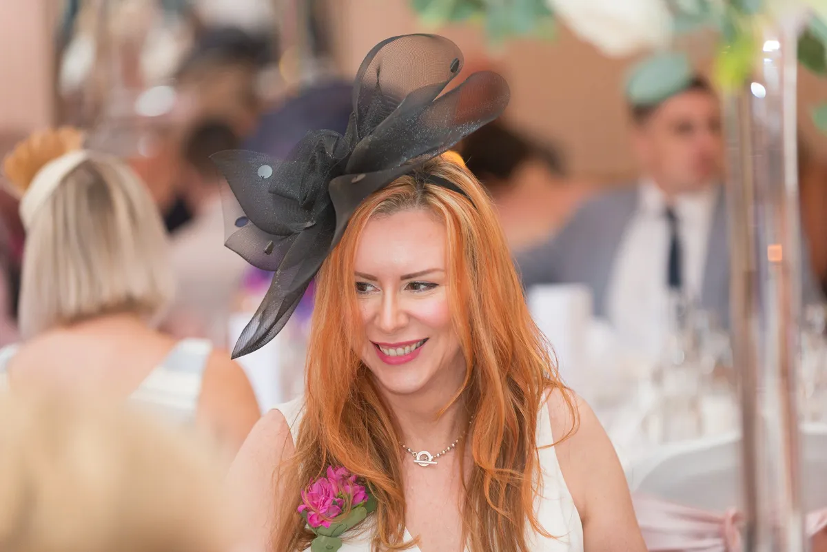 Smiling red-haired woman wearing a large black fascinator hat and a white dress with a pink flower corsage at a social event.