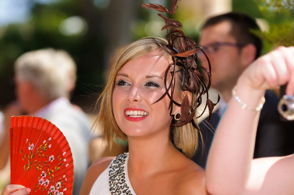 Smiling woman with short blonde hair wearing a decorative brown feathered headpiece and holding a red floral fan outdoors.