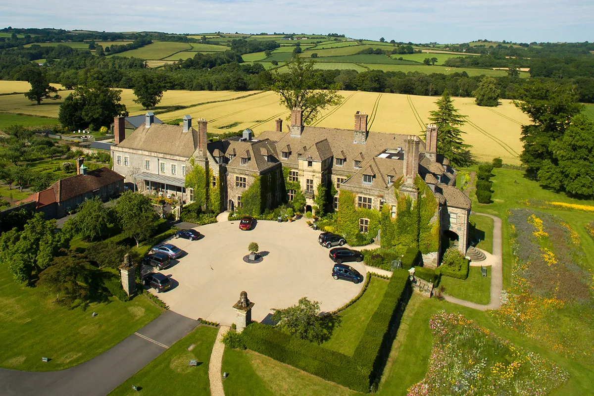 A large historic mansion with a circular driveway and several parked cars, surrounded by manicured lawns and farmland.