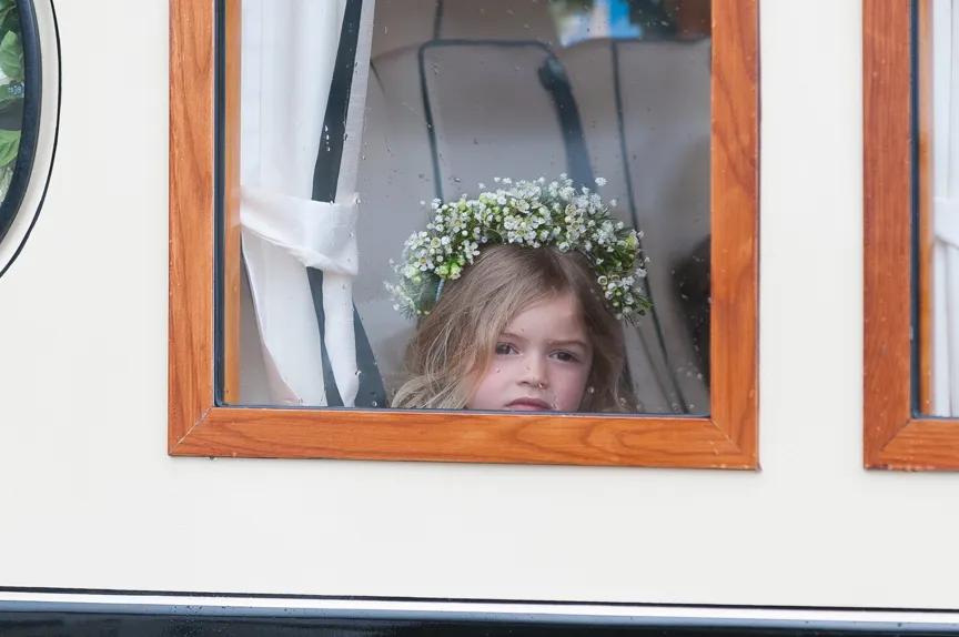 Young girl with a floral crown looking out of a wooden-framed window with white curtains.