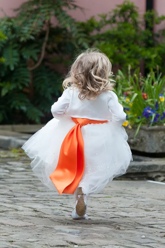 Young child with curly blonde hair wearing a white dress with an orange sash running on cobblestone path.