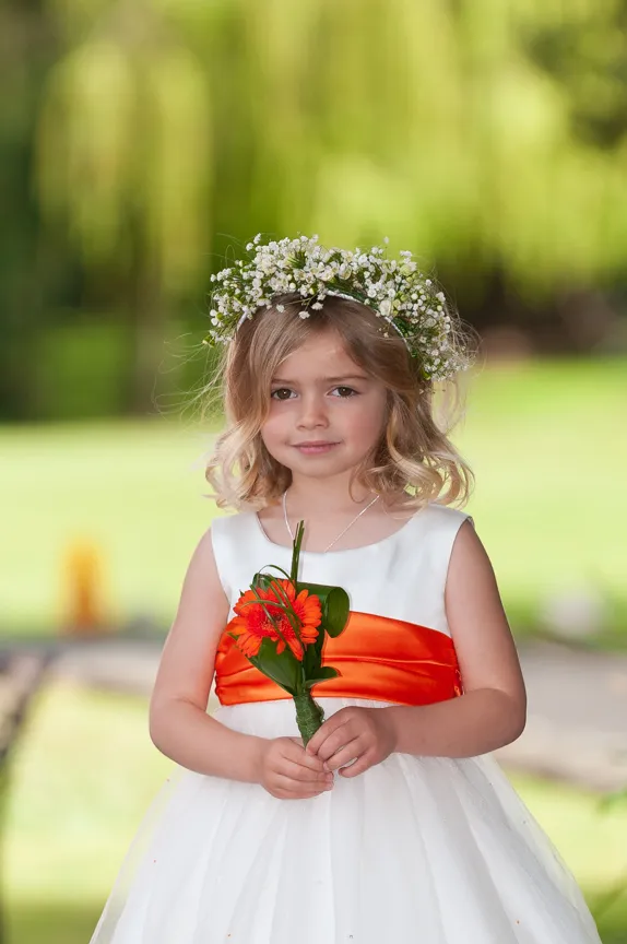Young girl with blonde hair wearing a white dress with an orange sash and a white flower crown, holding an orange flower bouquet.