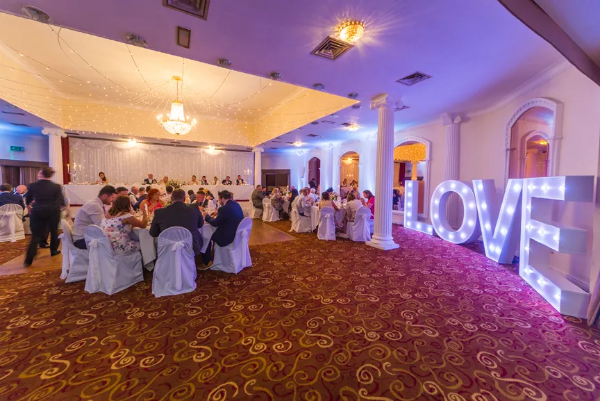Indoor wedding reception with guests seated at round tables covered in white cloths, and large illuminated letters spelling LOVE on the right.