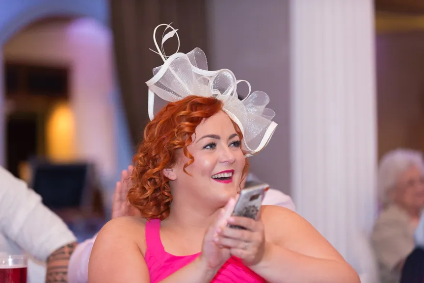 Smiling woman with curly red hair wearing a white decorative headpiece and a pink dress, holding a phone.