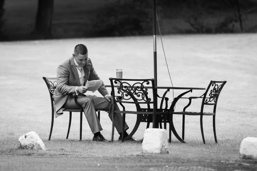 Man in a suit sitting alone at an outdoor metal table with chairs, reading a paper.