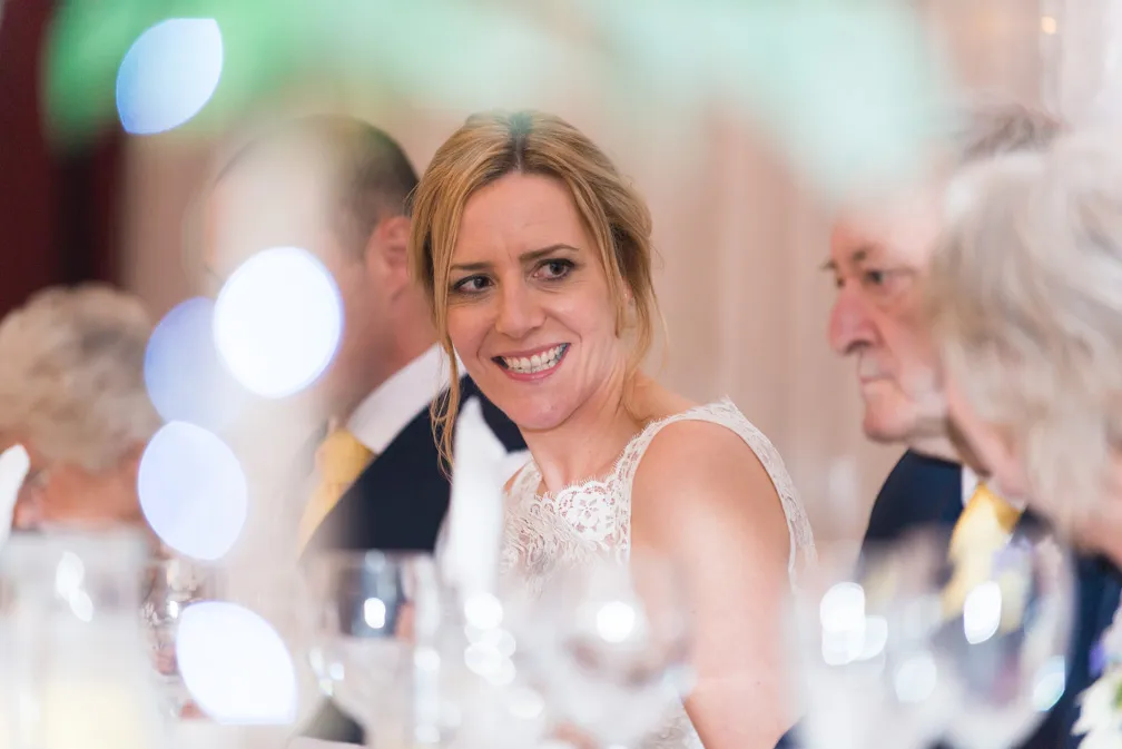 Smiling bride in a white lace dress sitting at a table with guests, blurred lights and glassware in the foreground.
