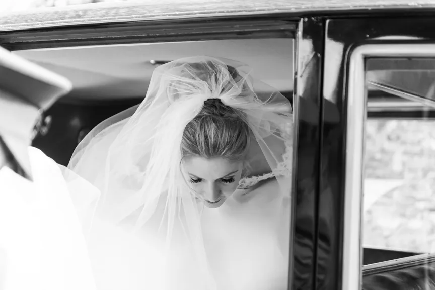Bride wearing a veil looking down as she sits inside a car.