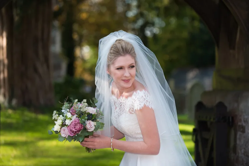 Bride in white lace wedding dress and long veil holding a bouquet of pink and white flowers outdoors near a stone archway.