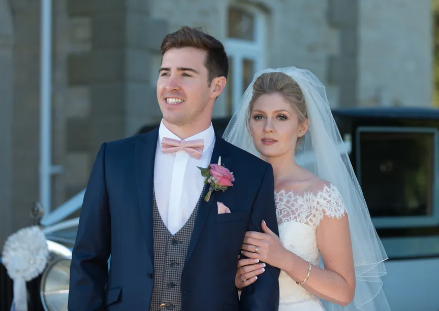 Wedding couple outdoors with the groom in a navy suit and pink bow tie and the bride in a white lace dress with a veil.