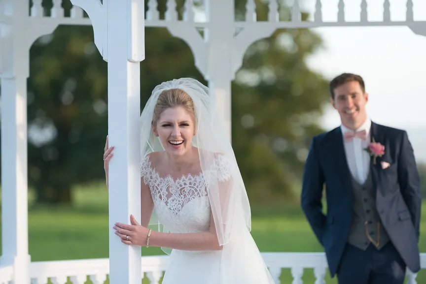 Bride in a lace wedding dress and veil smiling while leaning on a white gazebo post, with a groom in a suit and bow tie standing in the background outdoors.