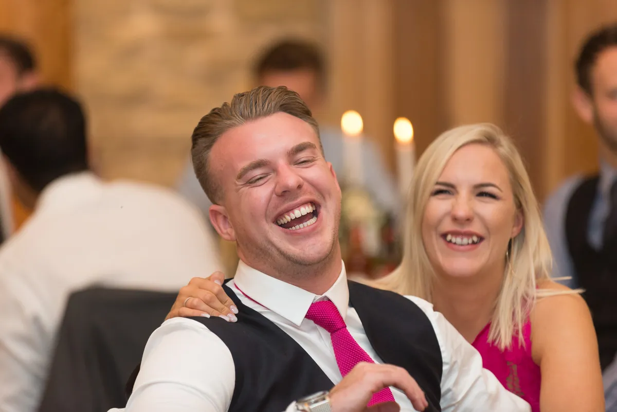 A man in a vest and bright pink tie laughing with a smiling woman in a pink dress behind him at an indoor event with candles in the background.