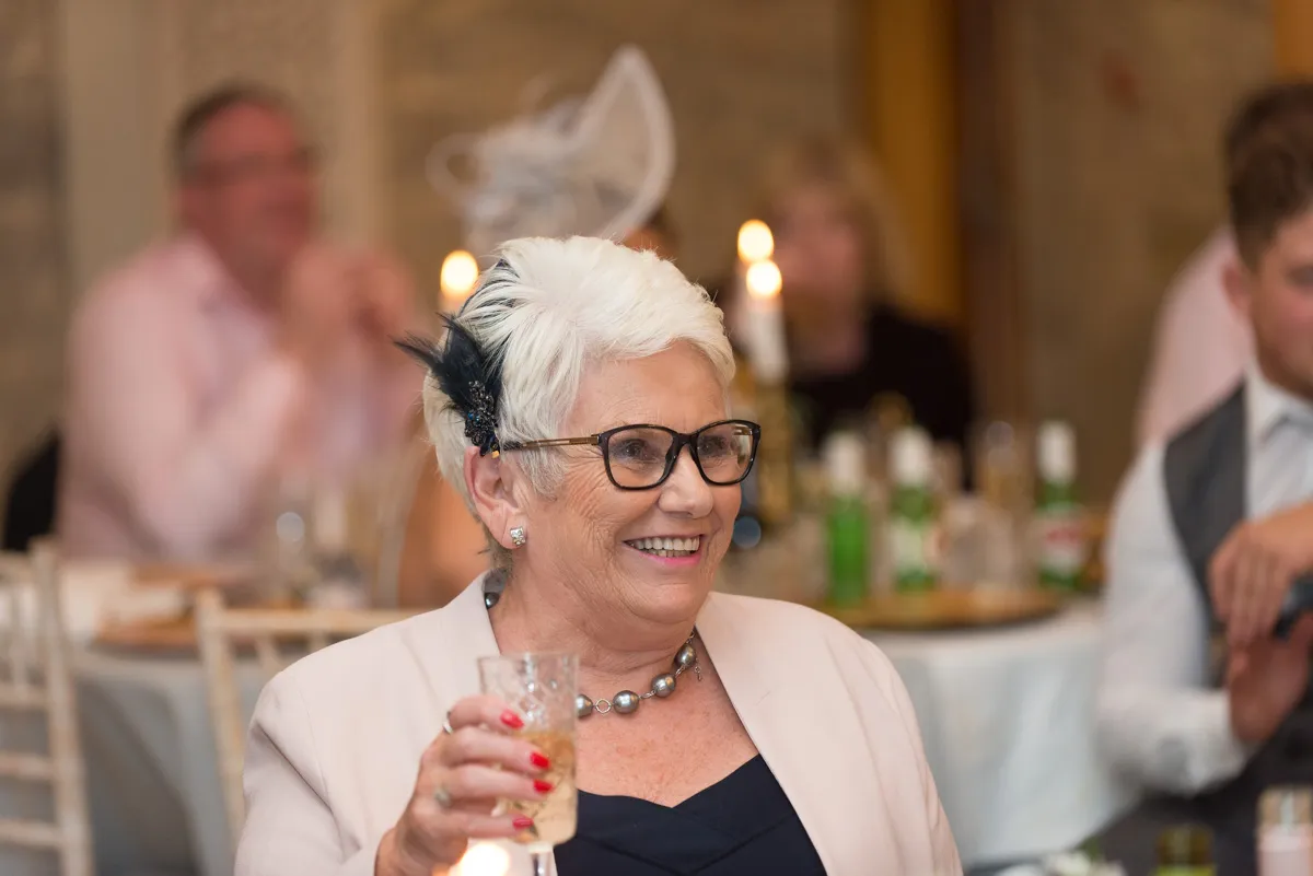 Elderly woman with short white hair, glasses, and a feather hair accessory smiling while holding a drink at a formal event.