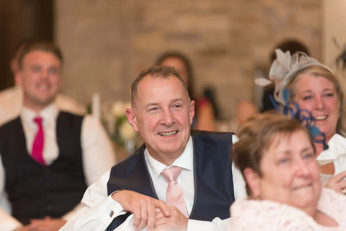 Smiling middle-aged man in a white shirt and pink tie with navy vest at a formal event, surrounded by other people.