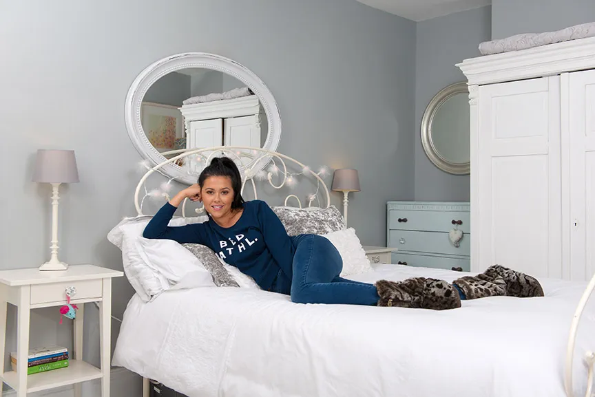 Woman in blue long-sleeve shirt and jeans lounging on a white bed in a cozy bedroom with gray walls.