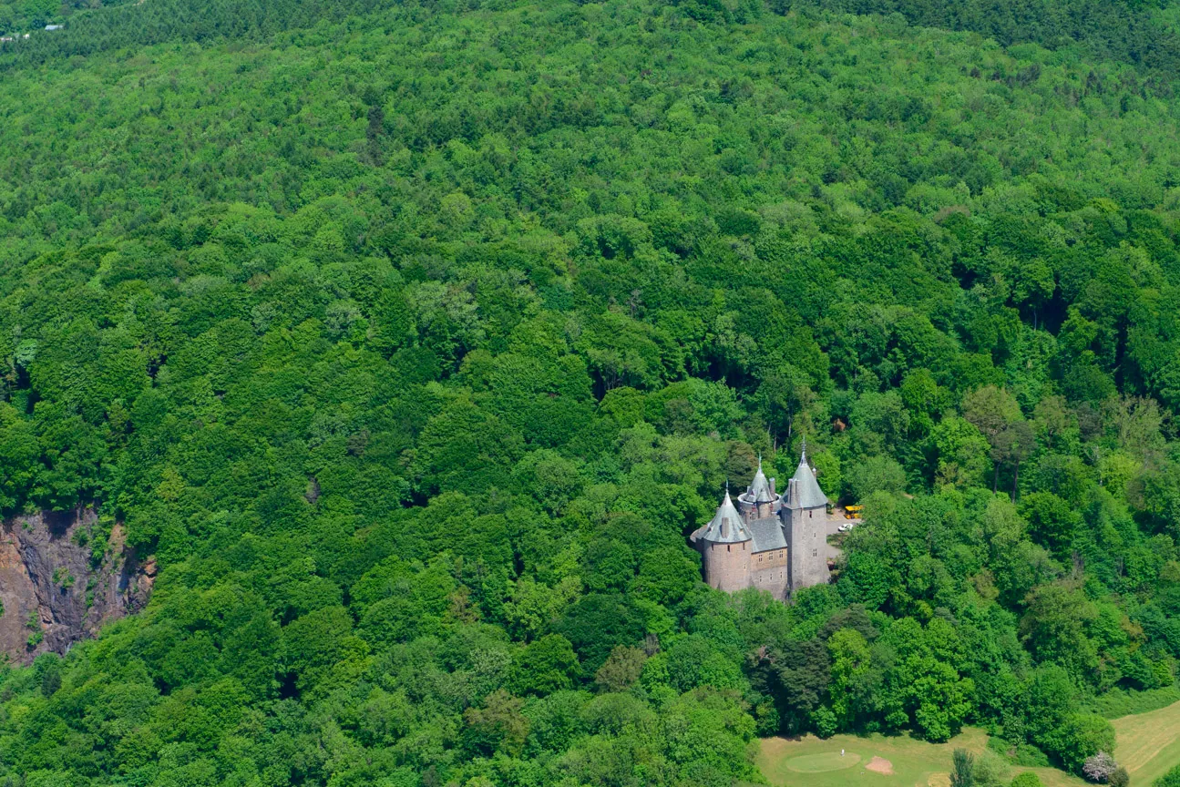 A castle with multiple towers nestled in dense green forest surrounded by rolling hills.