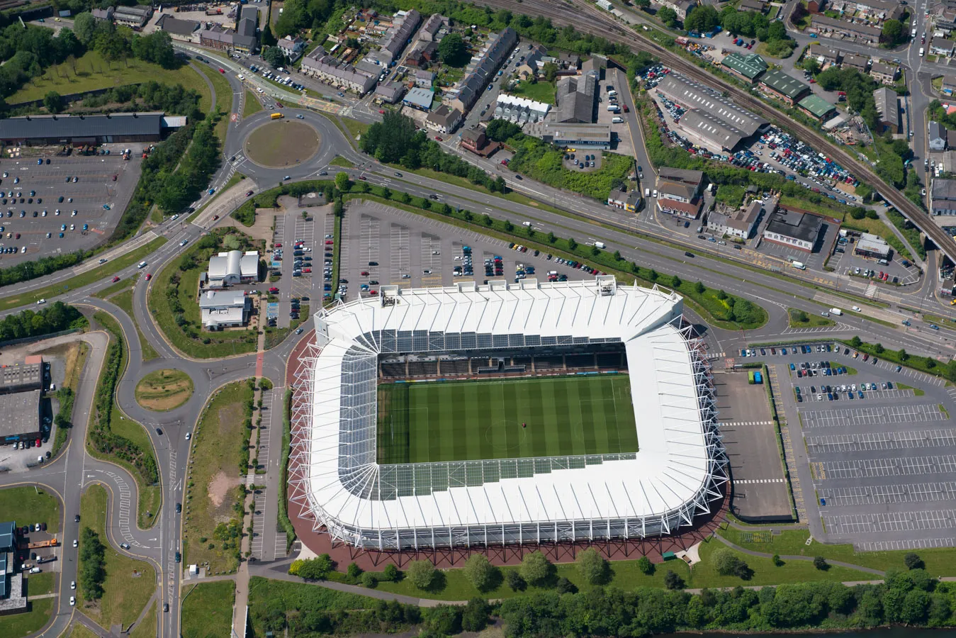 Aerial view of a large rectangular soccer stadium with a white roof surrounded by parking lots and roads.
