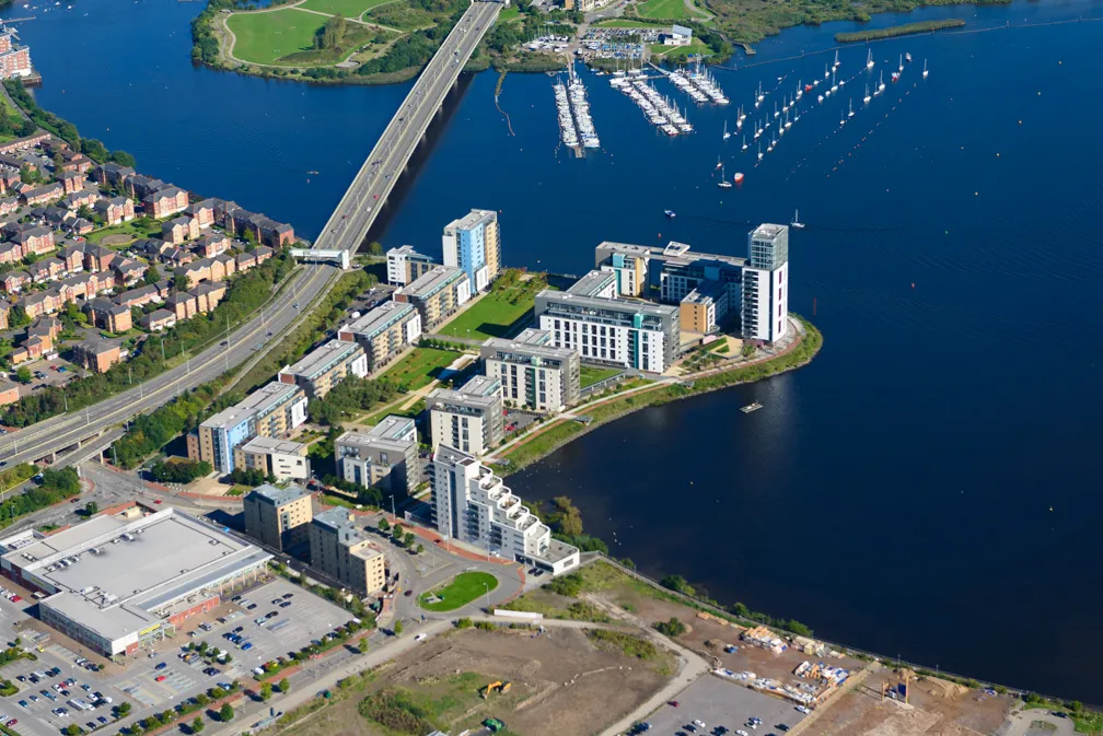 Aerial view of a waterfront residential area with modern apartment buildings, a marina with docked sailboats, and a nearby bridge over a body of water.