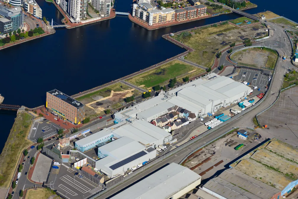 Aerial view of a large industrial complex with multiple white-roofed buildings next to a river and surrounded by roads and parking areas.