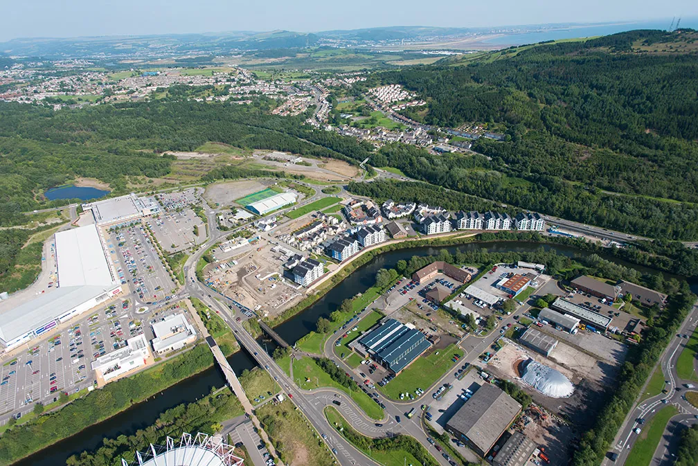 Aerial view of a town with residential buildings, industrial warehouses, parking lots, roads, lush green forests, and hills in the background.