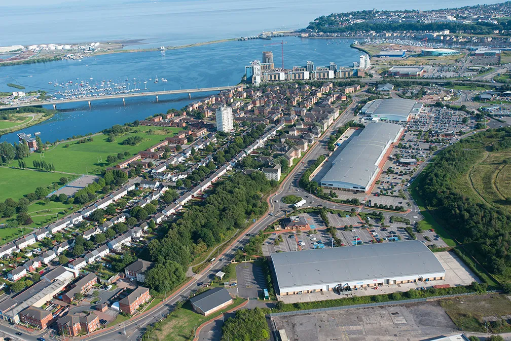 Aerial view of a coastal city area showing residential buildings, a river with a bridge, parking lots, and green spaces under a clear sky.