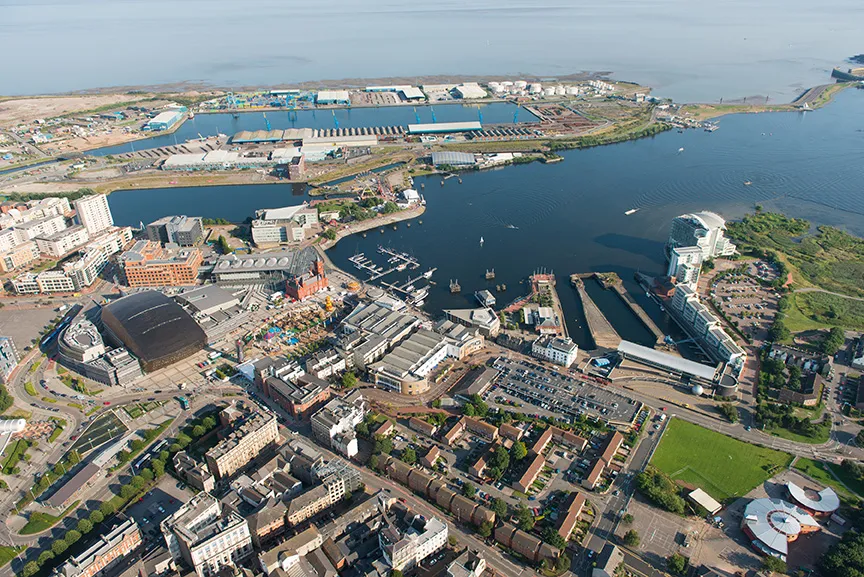 Aerial view of a coastal harbor area with boats docked, industrial tanks, urban buildings, roads, and green spaces.