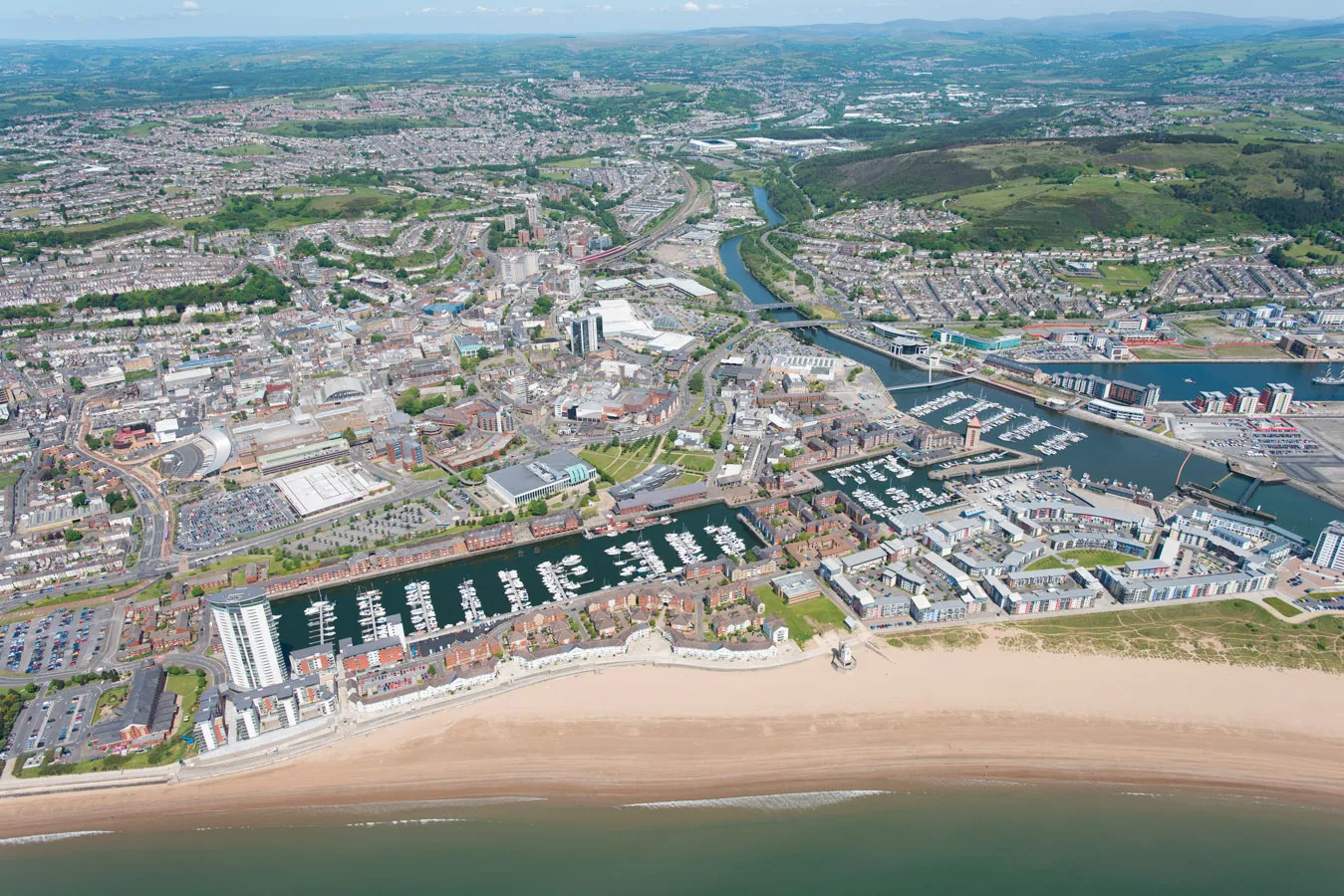 Aerial view of a coastal city featuring a marina with numerous boats, residential and commercial buildings, a sandy beach, and surrounding green hills.