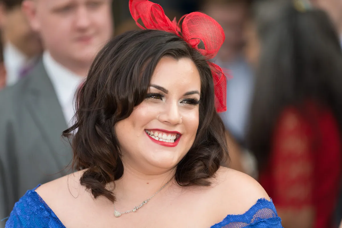 Woman with dark hair wearing red lipstick, a red bow headpiece, and a blue off-the-shoulder dress smiling.
