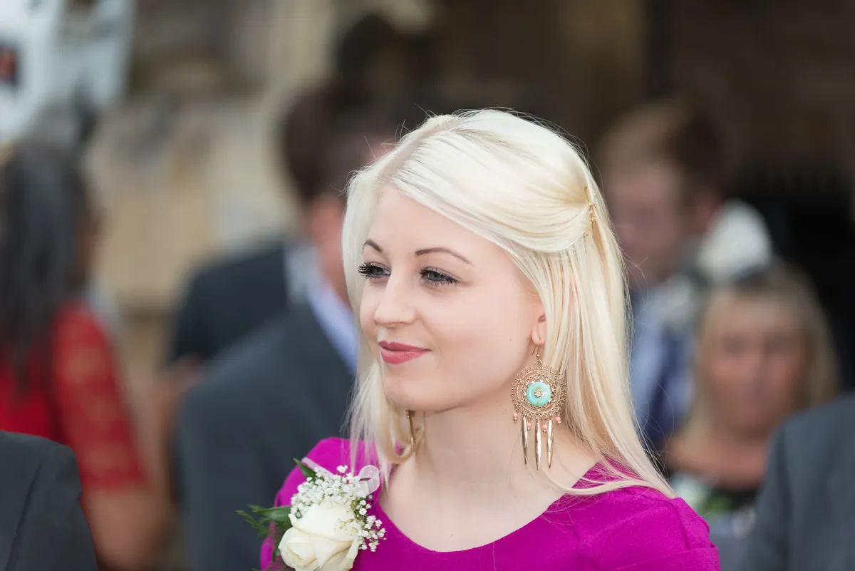 Young woman with long blonde hair wearing a purple dress and large decorative earrings, holding a white rose corsage.