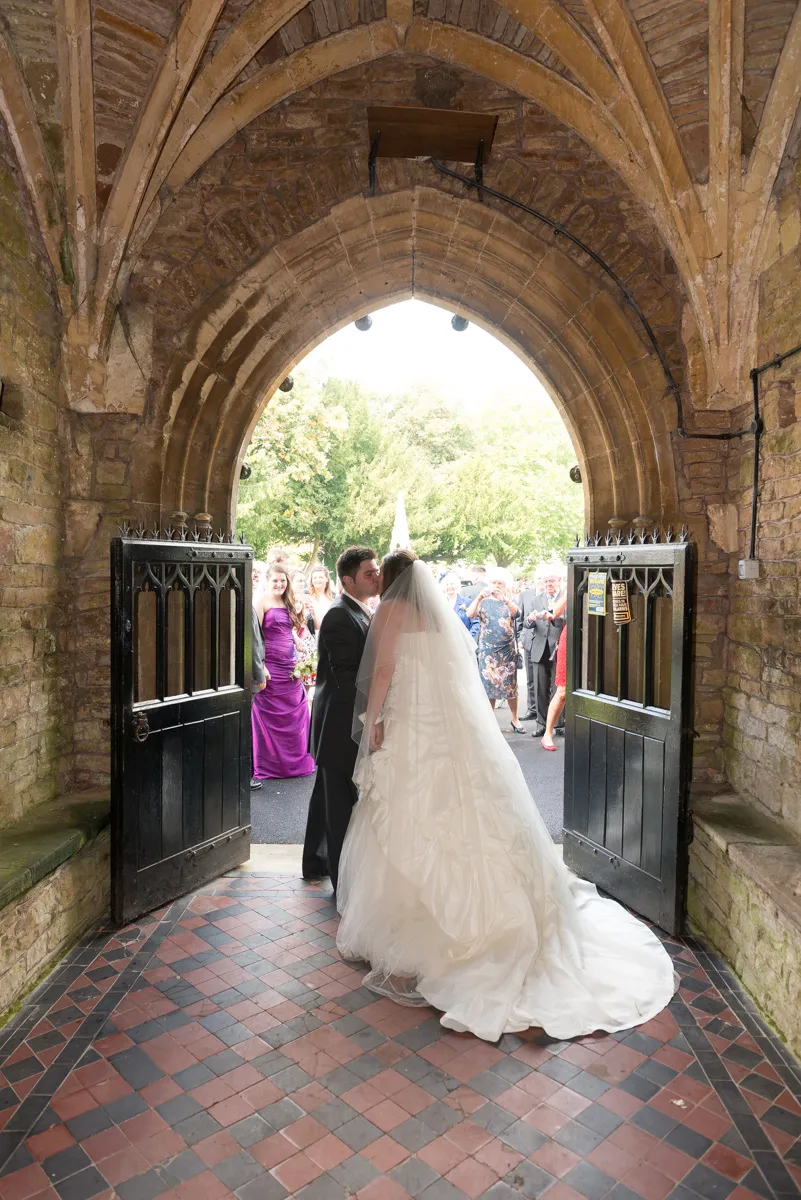 Newly married couple kissing under a stone archway with black open doors and wedding guests in the background.