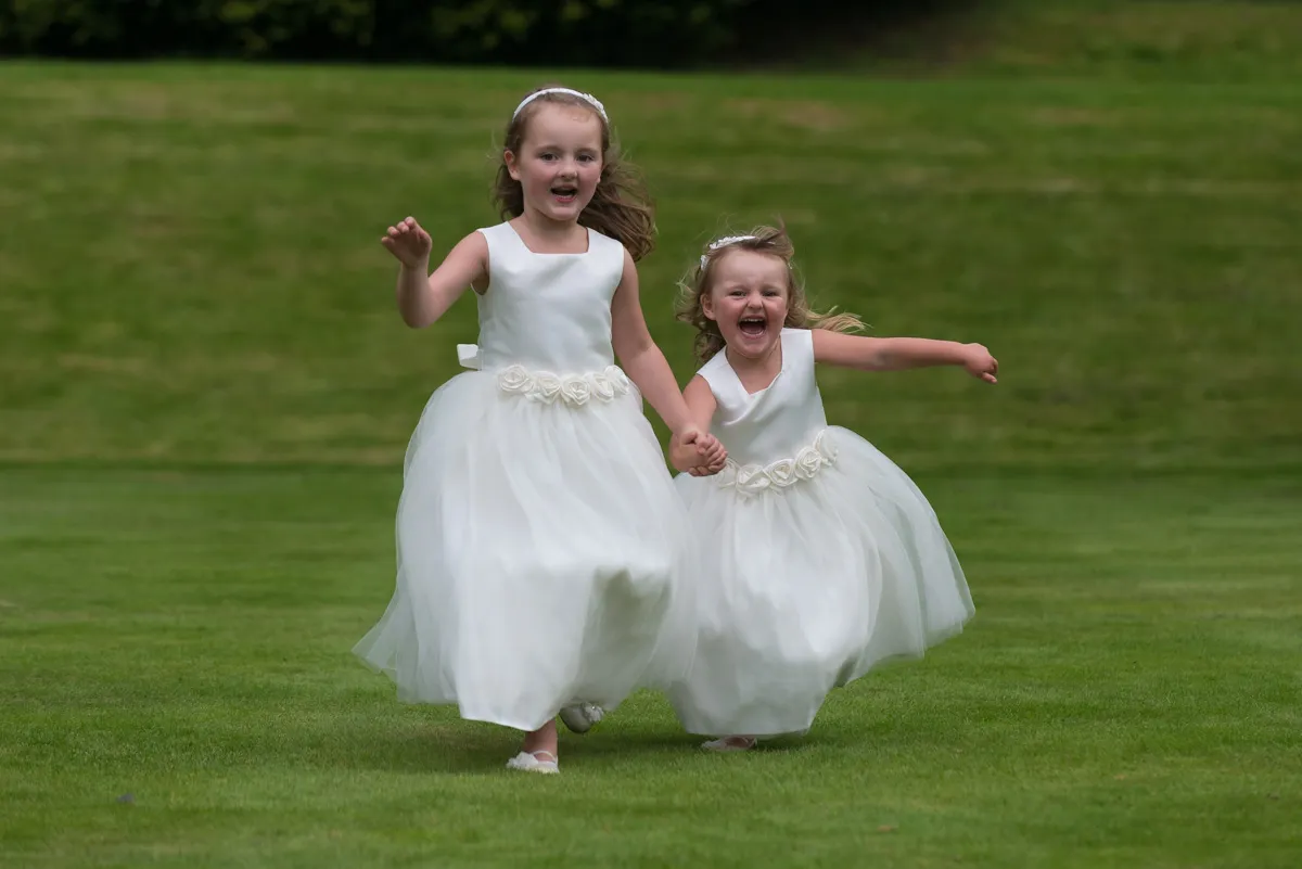 Two young girls in white dresses holding hands and running on green grass.