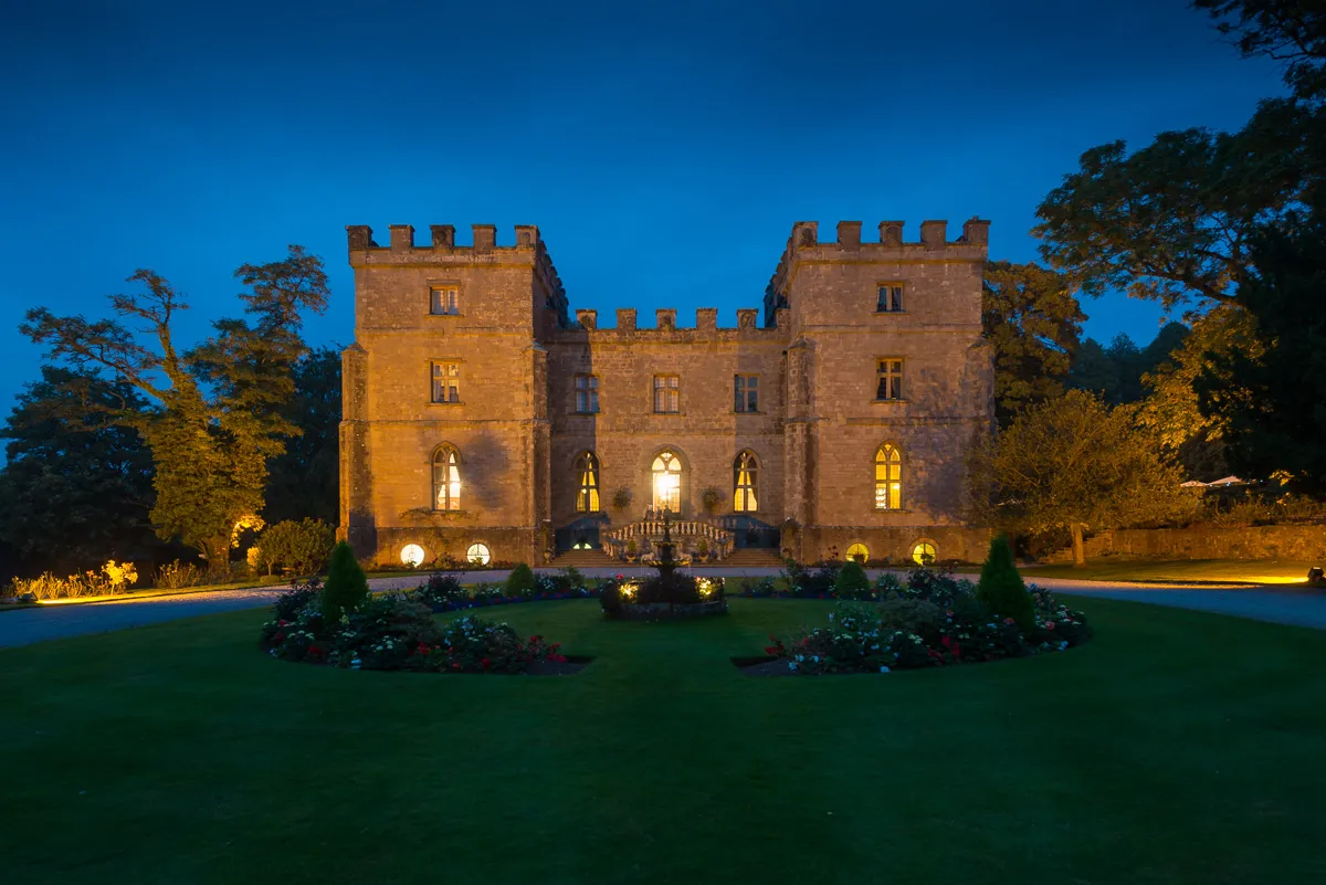 Stone castle illuminated at dusk with surrounding garden and trees under a deep blue sky.