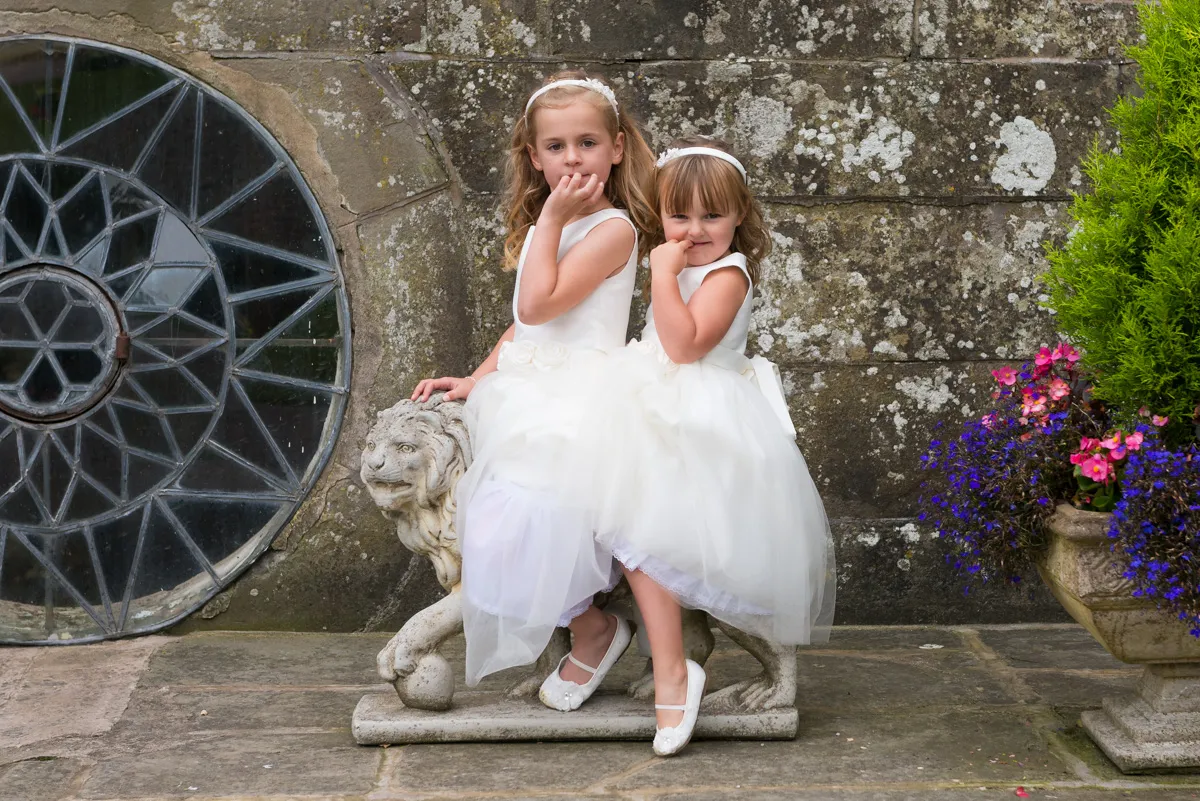 Two young girls in white dresses sitting on a stone lion statue against a weathered stone wall with a round window and colorful flowers nearby.