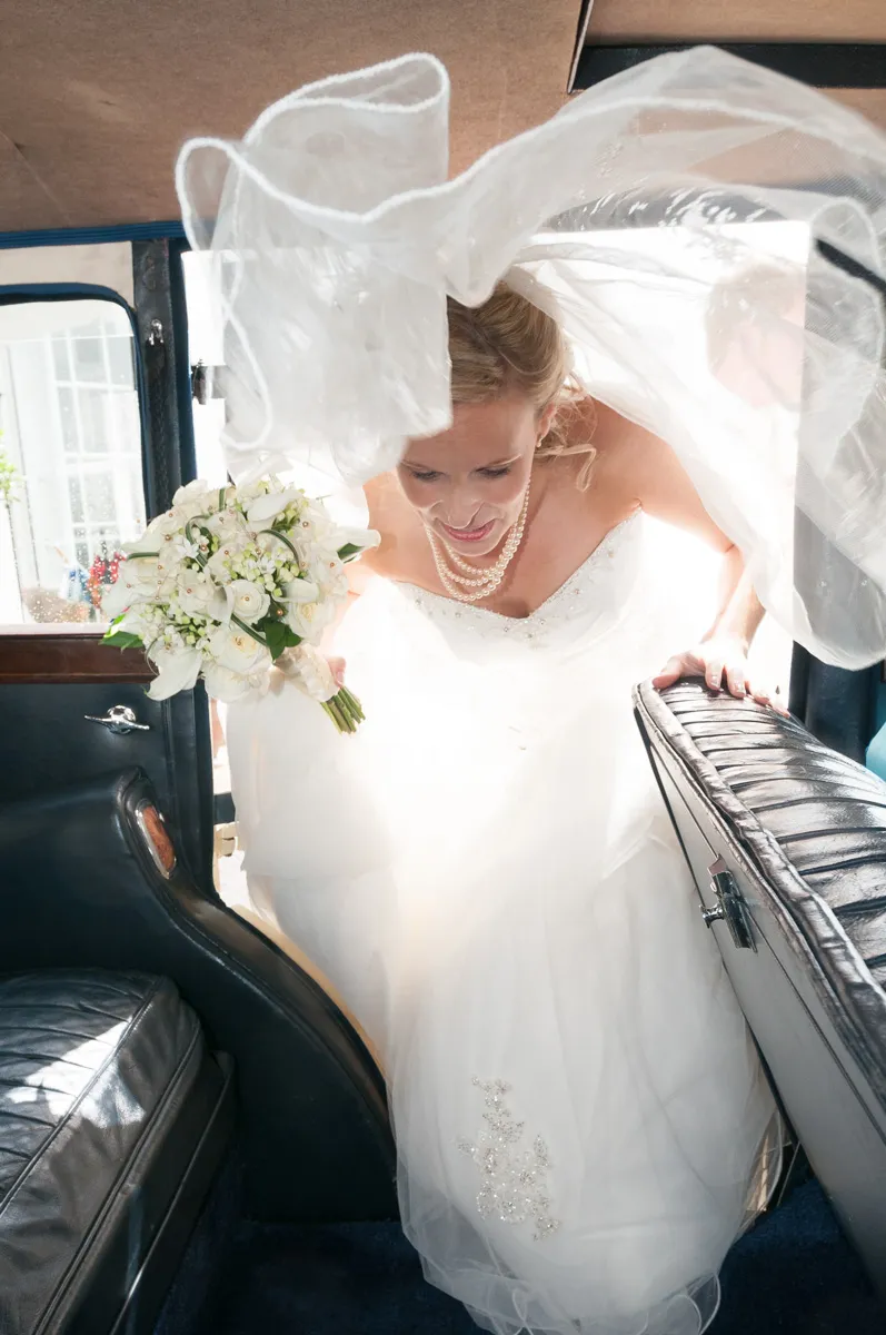 Bride in white wedding dress with veil and pearl necklace holding a bouquet of white flowers while stepping into a vintage car.