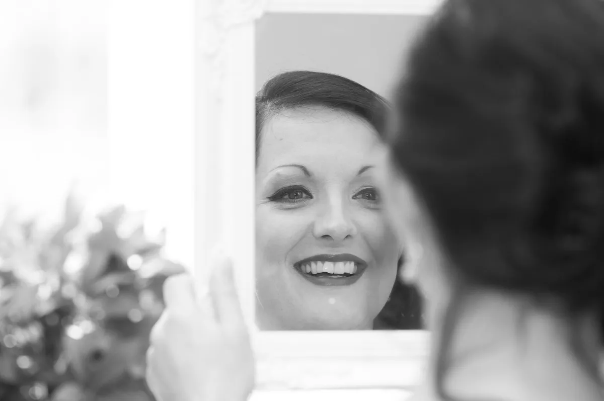 Black and white photo of a woman smiling at her reflection in a handheld mirror.