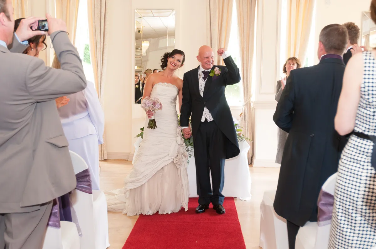 Bride and groom holding hands and smiling on a red carpet aisle in a wedding ceremony room.