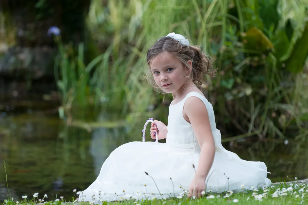 Young girl in a white dress sitting on grass beside a pond with wildflowers.