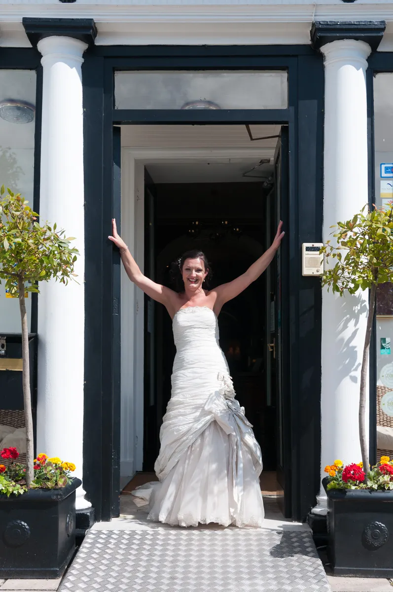 Bride in a strapless white wedding gown standing in a doorway with arms raised and smiling.