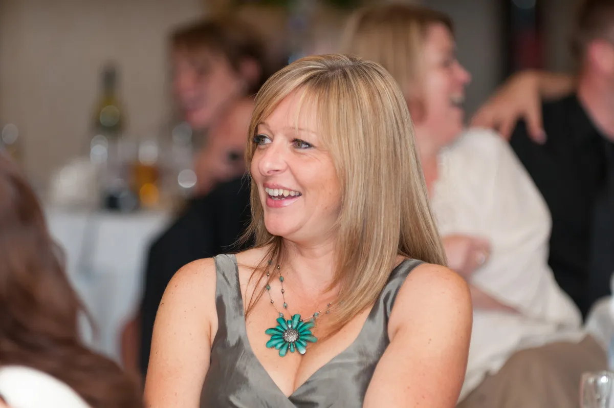 Smiling woman with blonde hair wearing a grey sleeveless dress and a turquoise flower necklace at a social event.