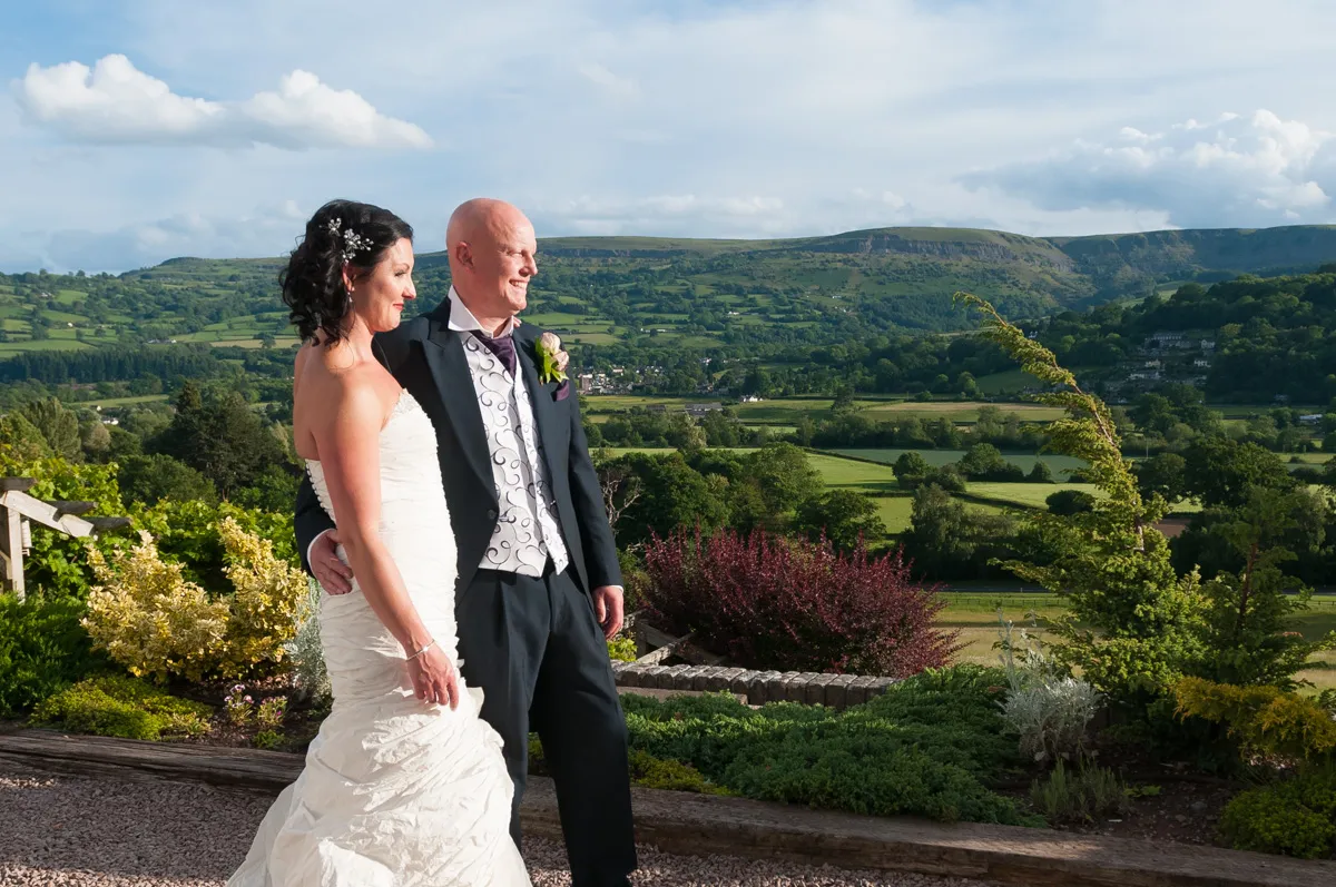 Bride in white dress and groom in dark suit standing together outdoors with a scenic green valley and hills in the background.