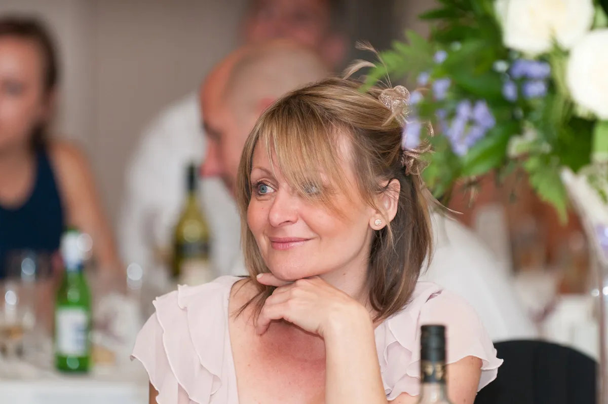 Smiling woman with shoulder-length hair resting her chin on her hand at a dining table with blurred background guests and flowers.