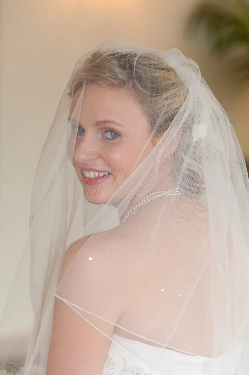 Bride with blonde hair wearing a pearl necklace and white veil, smiling over her shoulder.