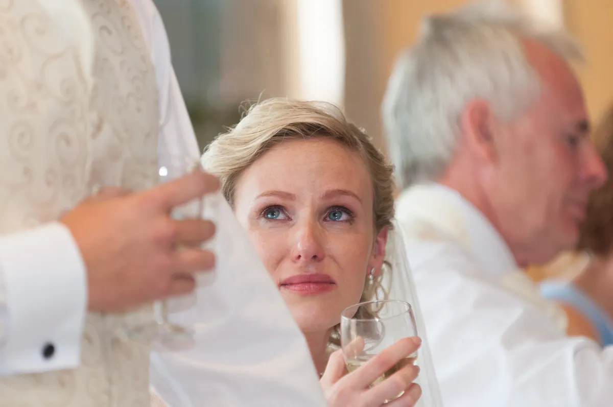 Woman holding a glass and looking up emotionally while a man, partially visible, in a cream vest gives a speech.