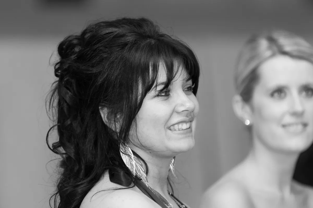 Smiling woman with long dark hair and feather earrings looking to the right, with a blurred woman in the background.