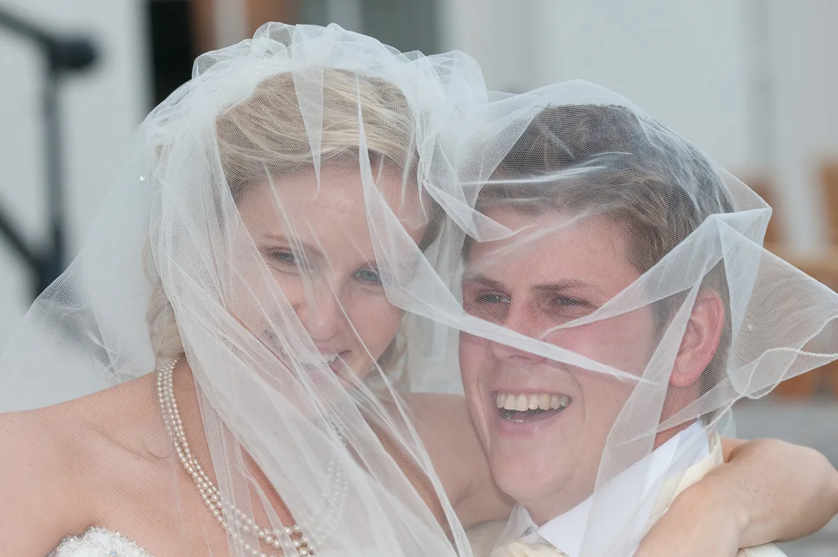 Bride and groom smiling happily with wedding veil over their faces.