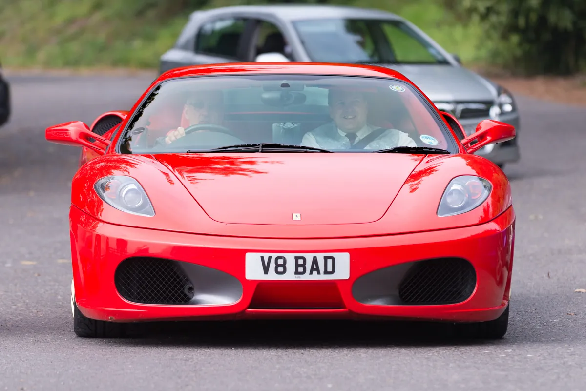 Front view of a red Ferrari sports car with license plate reading 'V8 BAD' driving on a road with two men inside.